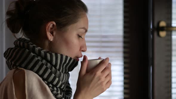 A sick woman warms up and drinks a hot drink near the window on a winter evening alt