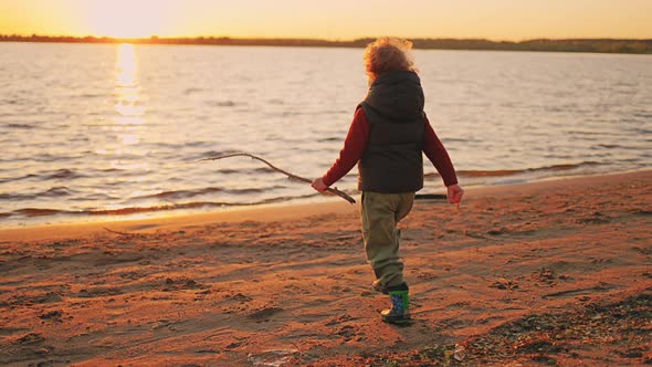 Cheerful Little Curly Boy is Running to Water of River in Sunset Time Walking on Sandy Coast in alt