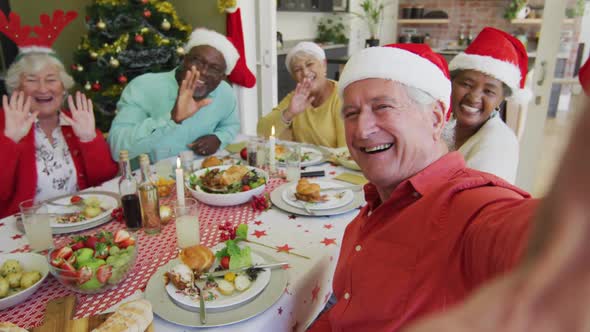 Happy diverse senior friends in santa hats taking selfie and waving at christmas dinner table alt
