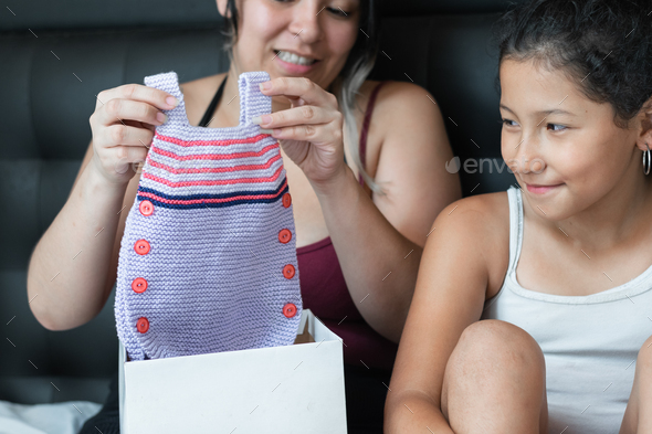 little brunette latina girl, next to her mom watching as her mom grabs the crocheted baby clothes.