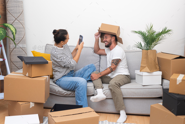 funny couple on moving day, unpacks belongings, having fun takin photo selfie on sofa in new home,