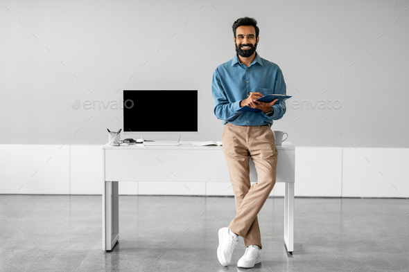 Happy indian male entrepreneur holding clipboard with document, standing near workplace in office