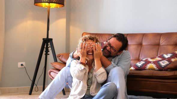 Adult man do a surprise to overjoyed woman at home sitting near the sofa - happy couple people enjoy alt