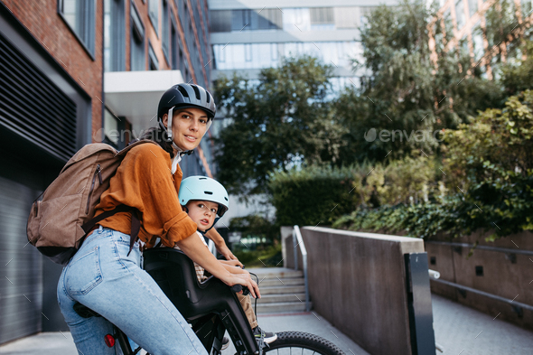 Mother carring her son on a child bike carrier, seat, both wearing helmets. Mom commuting with a