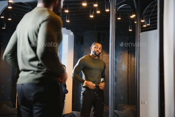 Stylish casual african american man at fitting room clothes store, looking on mirror.