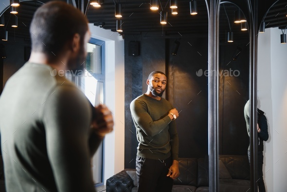 Stylish casual african american man at fitting room clothes store, looking on mirror.