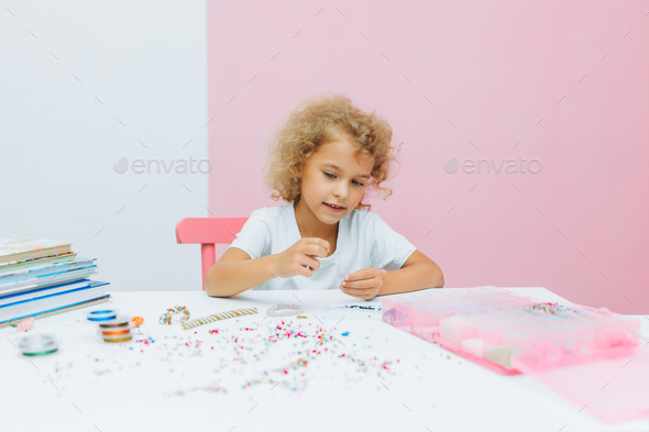 Cute little blonde girl making bead jewelry at a table in the room
