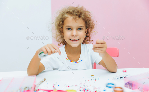 Cute little blonde girl making bead jewelry at a table in the room