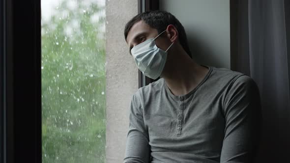 Depressed Man in Mask on Quarantine Looks at the Rain alt