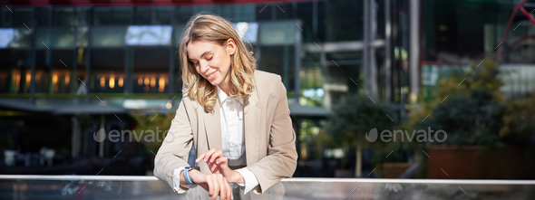 Smiling businesswoman looks at her digital watch, reads message or checks time, stands outside on