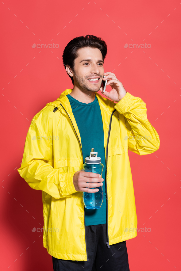 Smiling sportsman talking on smartphone and holding sports bottle on red background