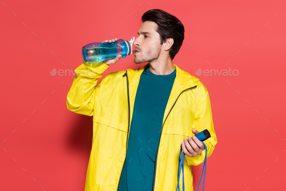 Brunette sportsman holding skipping rope and drinking water on red background