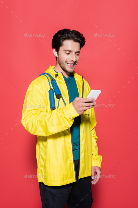 Sportsman in earphone and skipping rope on sports jacket holding smartphone on red background