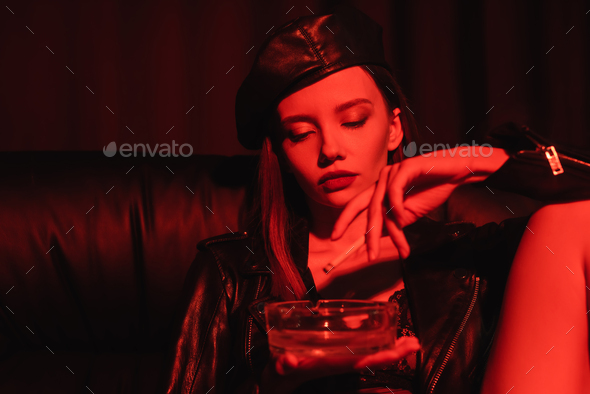 young sensual woman in leather jacket and beret smoking in red light on black background