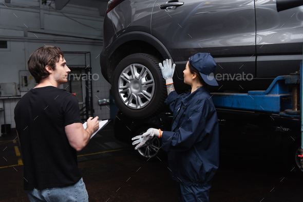 Mechanic pointing at car near colleague with clipboard in garage