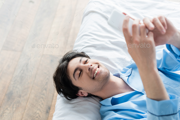 overhead view of happy man lying on bed and messaging on mobile phone