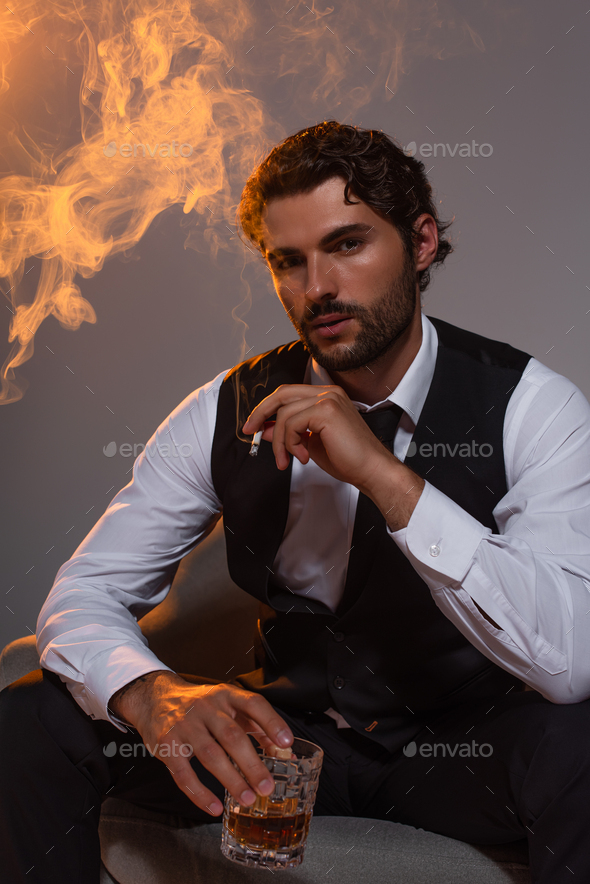 elegant man sitting in armchair with glass of whiskey and cigarette on ...