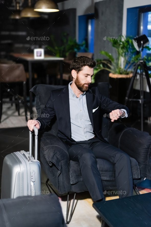 bearded man in suit holding luggage handle while checking time in hotel foyer