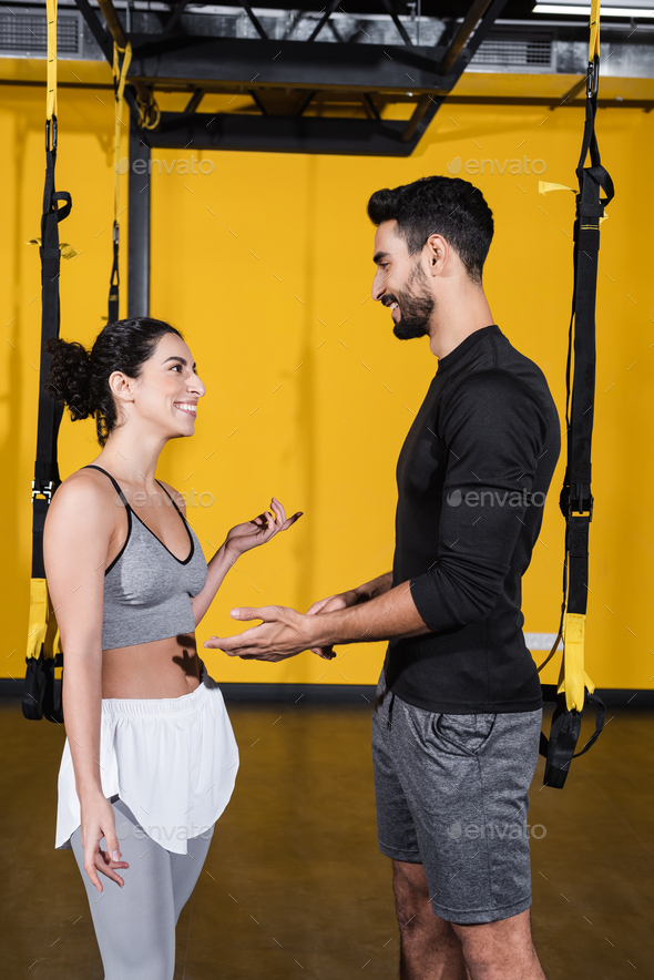Smiling arabian sportsman talking to middle east woman near suspension straps in gym