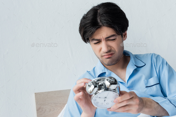 displeased man in blue pajamas holding vintage alarm clock in morning
