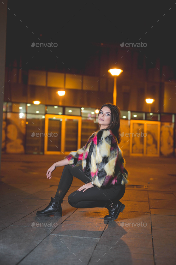 Vertical shot of a young female in a fashionable jacket posing squatted in the street at night