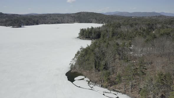 Drone shot flying forwards over a frozen lake with trees on the shores. alt