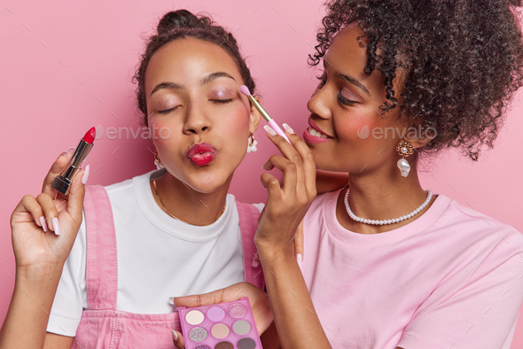 Photo of curly haired woman applies eyeshadow to friend do makeup together put on lipstick prepare