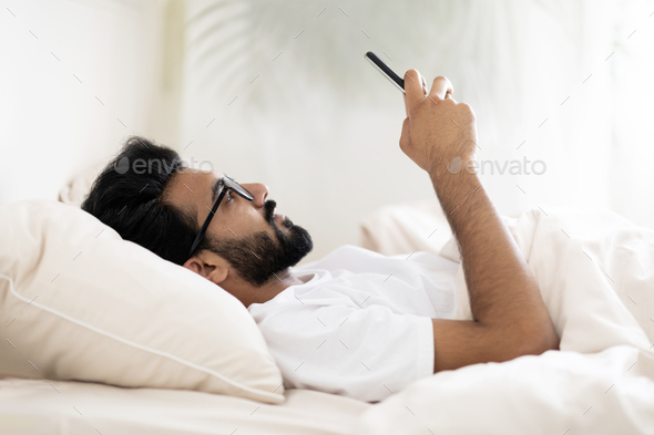 Gadget Addiction. Indian Man Browsing Internet On Smartphone While Lying In Bed