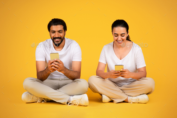 Smiling millennial middle eastern husband and wife chatting, typing on  phones, sit on floor Stock Photo by Prostock-studio