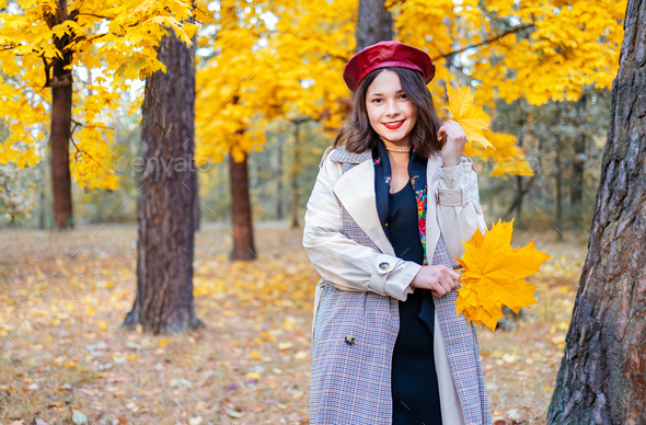 Hello october. happy trendy woman in red hat and beige coat rejoicing in the city park at sunset.