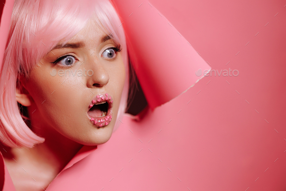 Surprised face of a girl model with bright makeup looks into a hole in pink paper