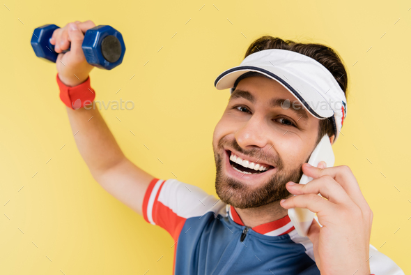 Smiling sportsman talking on smartphone and looking at camera while training with dumbbell isolated