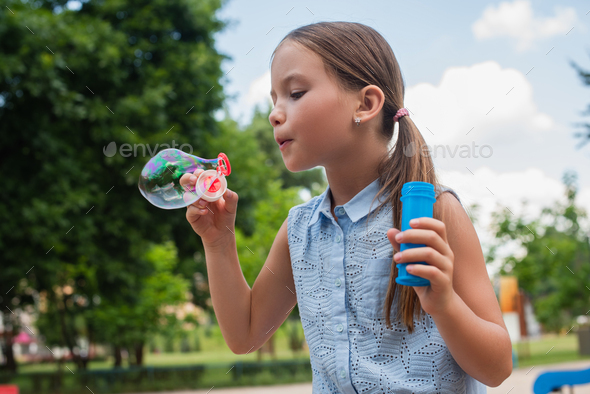 girl in sleeveless blouse blowing soap bubble in park