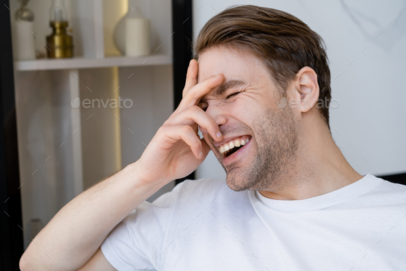 excited man in white t-shirt obscuring face with hand while laughing at home