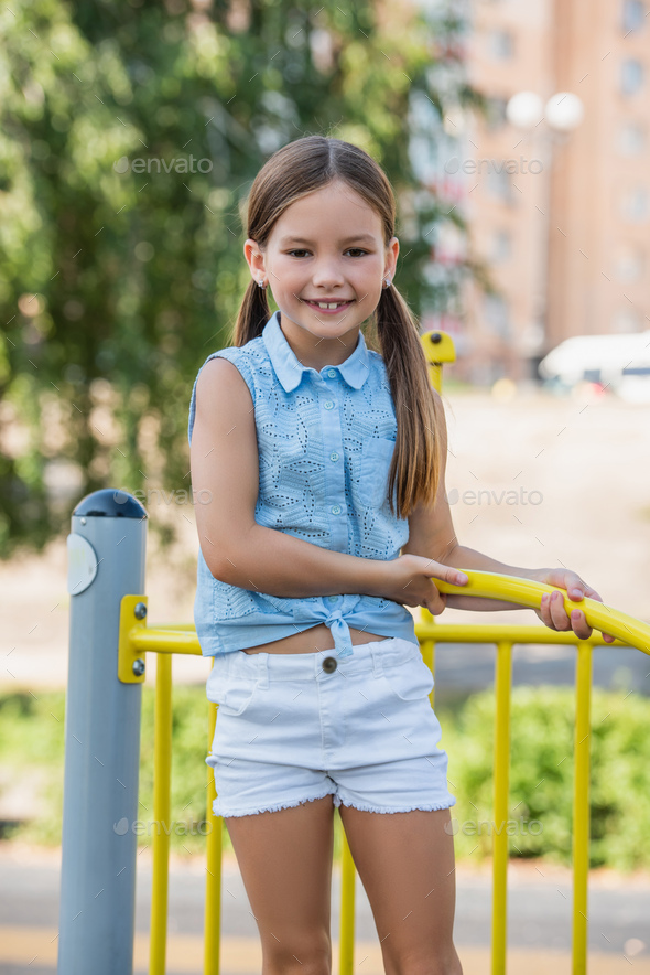 joyful girl in sleeveless blouse and short smiling at camera on playground