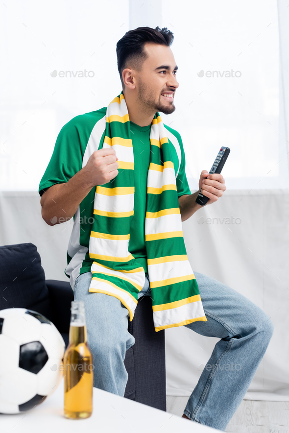 thrilled man in striped scarf holding clenched fist for luck while watching football match at home