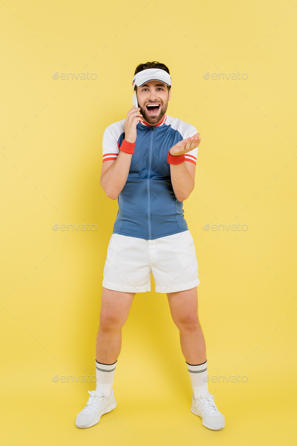 Full length of excited sportsman talking on smartphone and looking at camera on yellow background