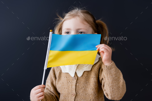 little girl obscuring face with small ukrainian flag isolated on black