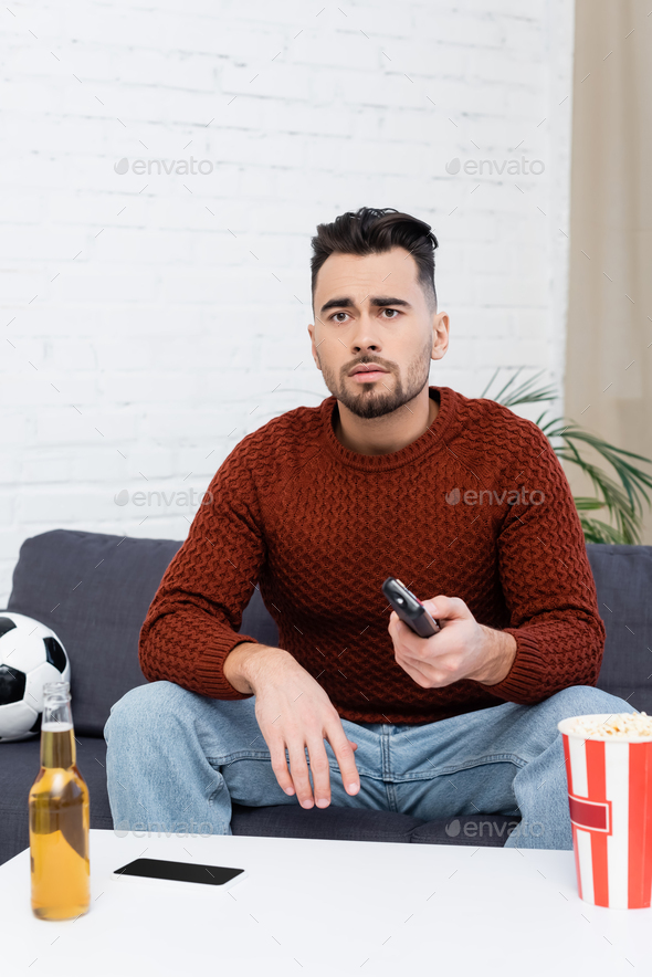 sad sports fan with remote controller watching game on tv near soccer ball, beer and popcorn
