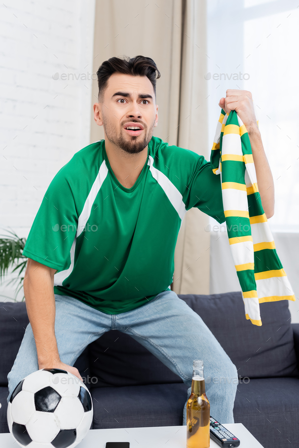 worried and excited sports fan holding striped scarf while watching football match on tv
