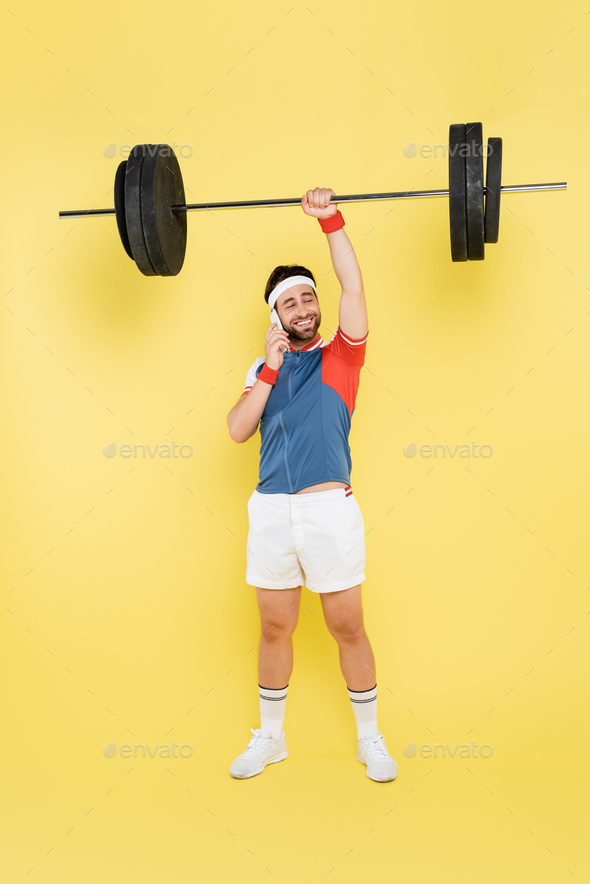Full length of smiling sportsman talking on cellphone and lifting barbell on yellow background