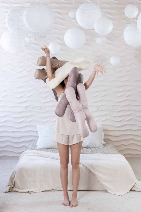 Girl in beige pajamas throws up a stack of silk beauty pillows on the  background of a bed. Vertical Stock Photo by Tatiana_Mara