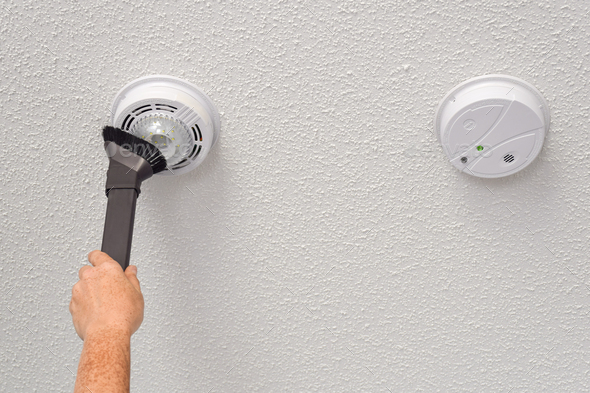 Person\'s hand vacuuming smoke and carbon monoxide detectors on a ceiling of a domestic room