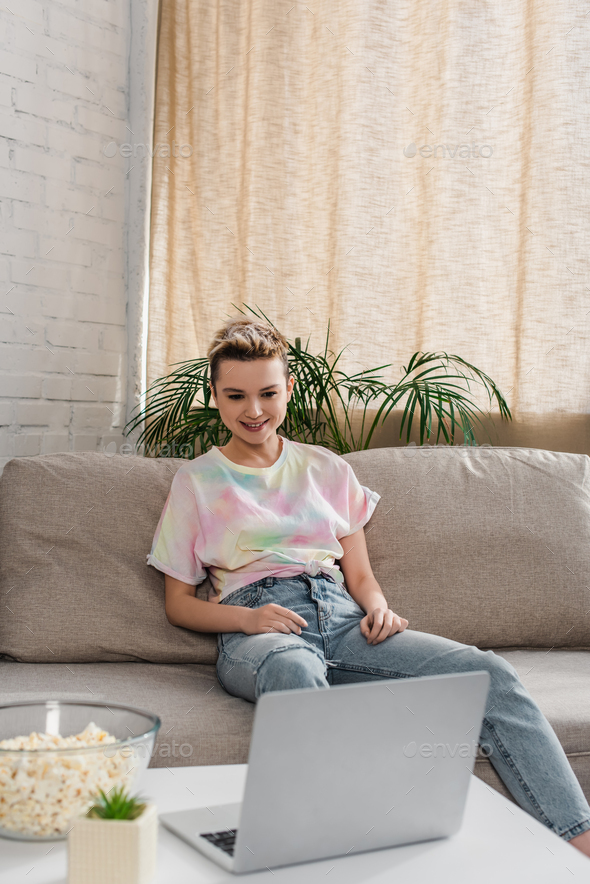 young and happy pansexual person with short hair sitting on couch and watching film on laptop