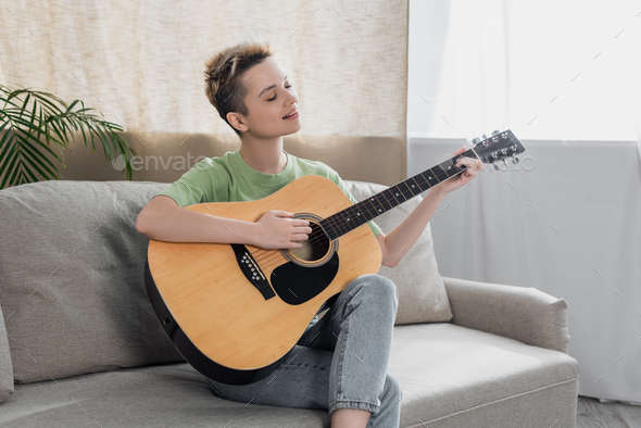 smiling bigender person with short hair sitting on couch and playing acoustic guitar