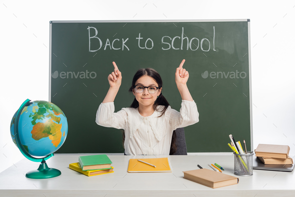 Smiling schoolkid pointing at chalkboard with back to school lettering near globe and books isolated