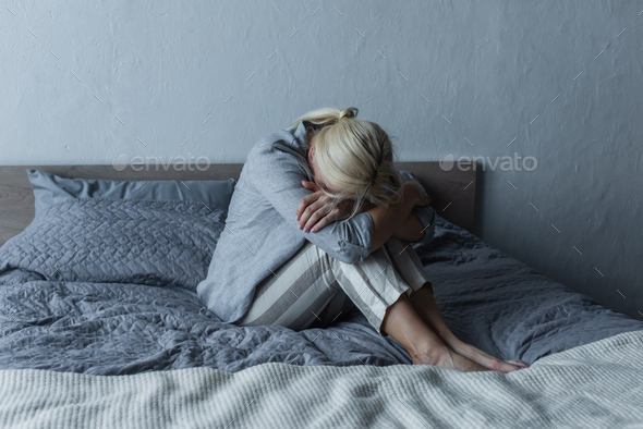 blonde woman covering face and hugging knees on bed while feeling pain in stomach during menopause