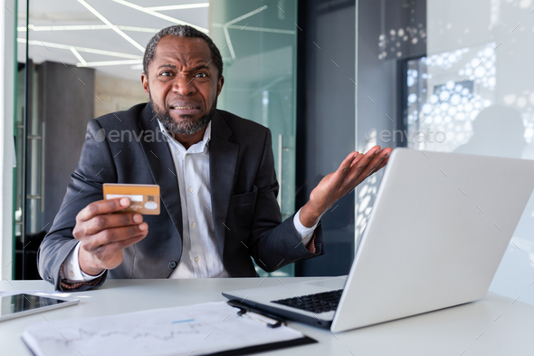 Portrait of upset cheated businessman inside office, mature boss holding bank credit card in hands