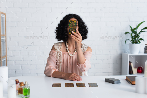 KYIV, UKRAINE - JUNE 29, 2022: brunette astrologer obscuring face with tarot card during spiritual