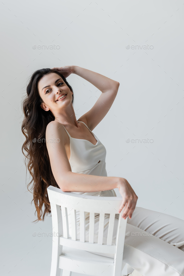 Positive Long Haired Woman Sitting On White Chair Isolated On Grey